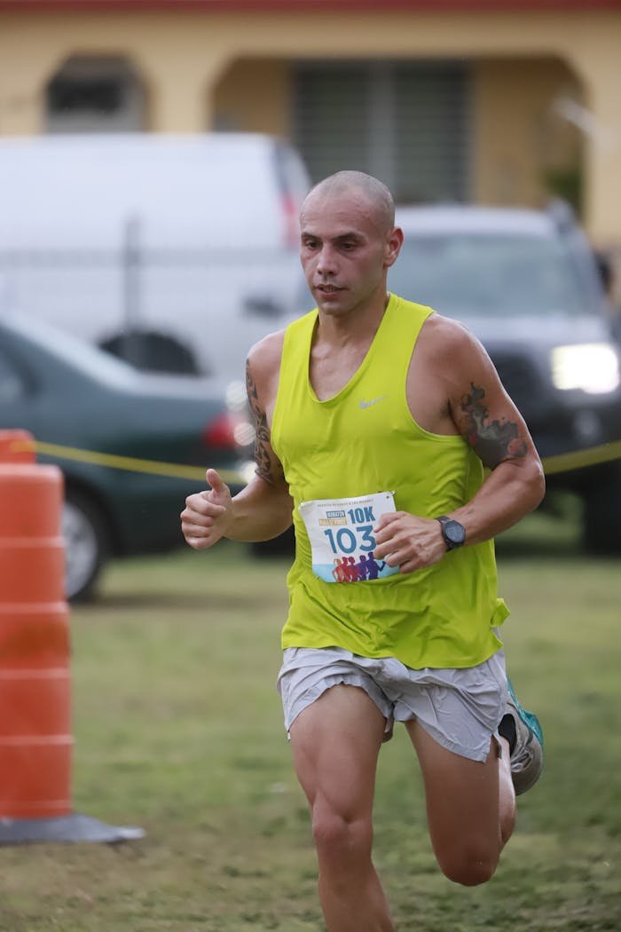 about-us-01 Athletic male competing in a 10K race, wearing a green tank top on a grassy outdoor track.